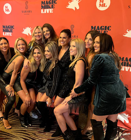Group of women in sparkly dresses and boots smiling and posing together on a red carpet backdrop during a Las Vegas girls' night out.