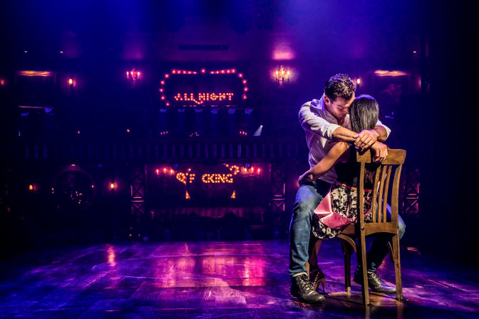 Romantic couple embracing on a wooden chair center stage in a hazy cabaret-style theater, purple-pink spotlights and vintage marquee bulbs reading “ALL NIGHT” in the background