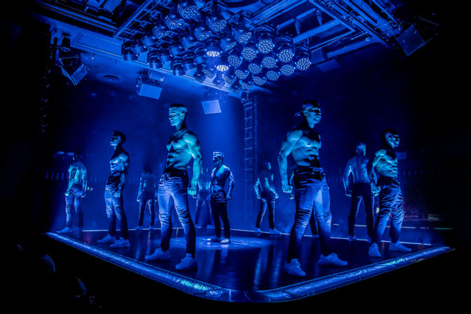 Shirtless male dancers striking poses on an illuminated stage under intense blue LED stage lighting during a high-energy nightclub performance