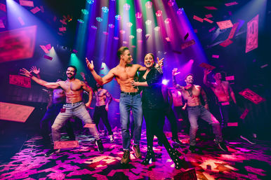 High-energy nightclub stage with a woman in a sparkly jacket dancing alongside shirtless male performers under colorful spotlights as falling dollar bills rain down.