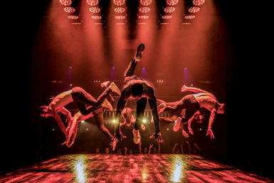 High-energy acrobatic dance troupe of shirtless performers mid-air on a wooden stage under dramatic red spotlights, audience silhouettes in an indoor theater.