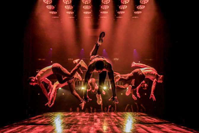 High-energy acrobatic dance troupe of shirtless performers mid-air on a wooden stage under dramatic red spotlights, audience silhouettes in an indoor theater.