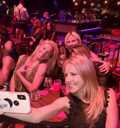 Group of smiling women taking a selfie at a lively piano bar lounge with cocktails, wooden tables and warm red-pink stage lighting — nightlife scene