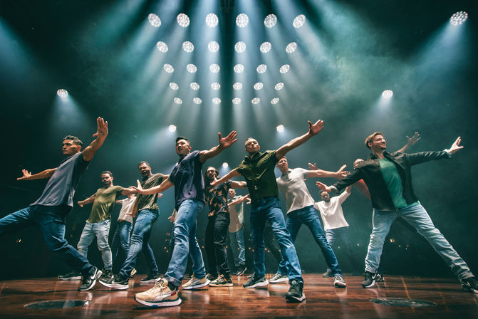 Energetic group of dancers on a wooden stage striking a synchronized wide‑arm pose beneath dramatic circular spotlights and smoky stage lighting — live performance