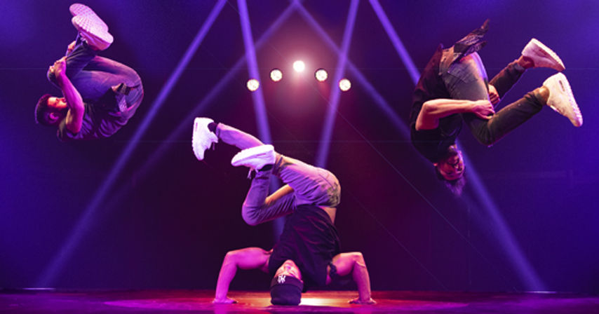 Three breakdancers — center freeze/headstand with two dancers executing mid-air flips on either side — performing on an indoor stage bathed in purple spotlights and round backlights, wearing casual streetwear and white sneakers.
