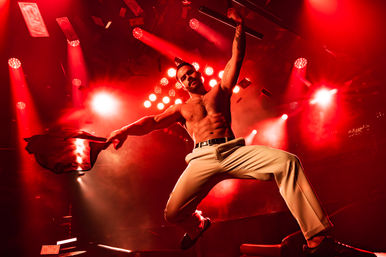 Muscular shirtless performer mid-jump on a red-lit nightclub stage, waving a jacket as spotlights blaze and banknotes flutter through the air.