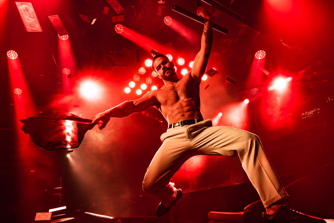 Muscular shirtless performer mid-jump on a red-lit nightclub stage, waving a jacket as spotlights blaze and banknotes flutter through the air.
