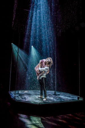 Two performers in a contemporary dance embrace on a rain-drenched theater stage, lifted amid spotlight beams and cascading water.