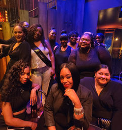 Group of women enjoying an urban nightlife lounge, gathered around a woman in a sparkly outfit wearing a celebratory sash — festive birthday/party vibe with warm purple-blue lighting.