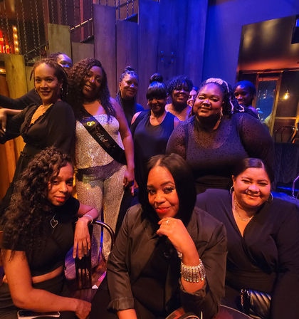Group of women enjoying an urban nightlife lounge, gathered around a woman in a sparkly outfit wearing a celebratory sash — festive birthday/party vibe with warm purple-blue lighting.