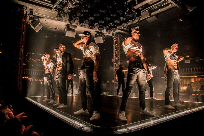 Energetic male dancers performing a choreographed late-night nightclub stage show on an illuminated platform with dramatic overhead lights and a nearby audience.