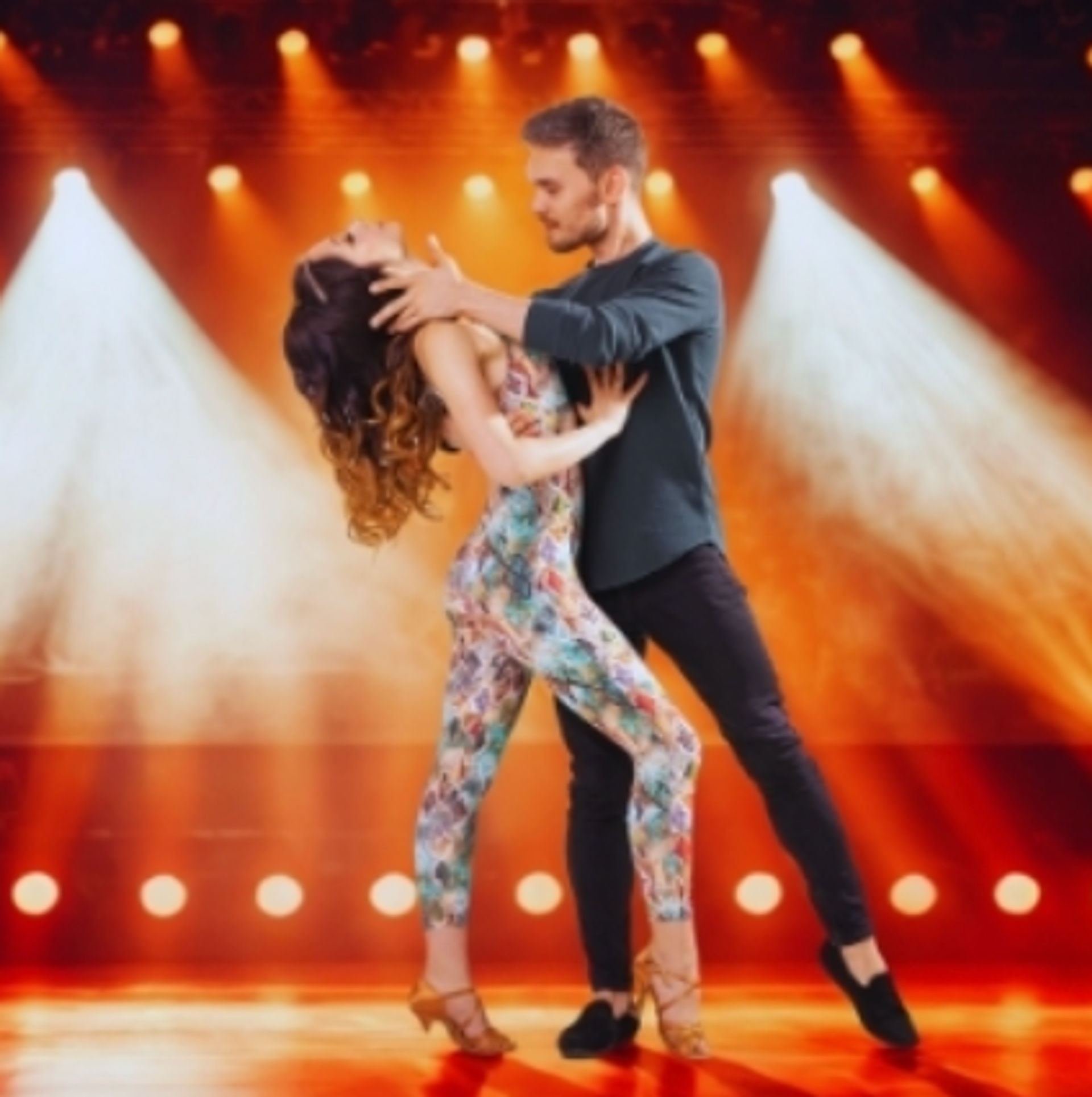Couple performing a dramatic partner dance on a theater stage under warm spotlights, woman in a floral jumpsuit and heels.