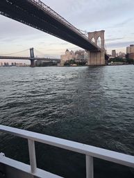 Boat-level view under the Brooklyn Bridge over the East River in New York City, with the Manhattan Bridge and Manhattan skyline at dusk.