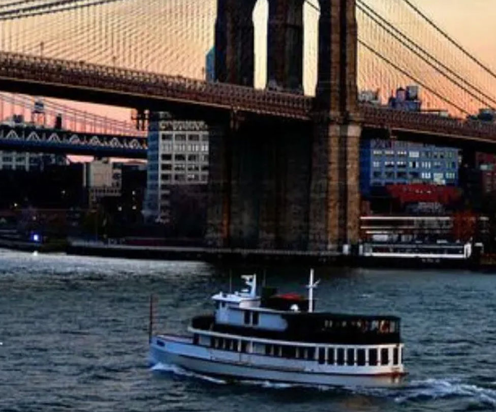 Sightseeing ferry chugging along the East River under the Brooklyn Bridge at sunset with Manhattan skyline in the background
