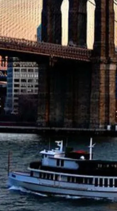 White sightseeing ferry cruising on the East River beneath the Brooklyn Bridge at sunset with city skyline and another suspension bridge in the background.