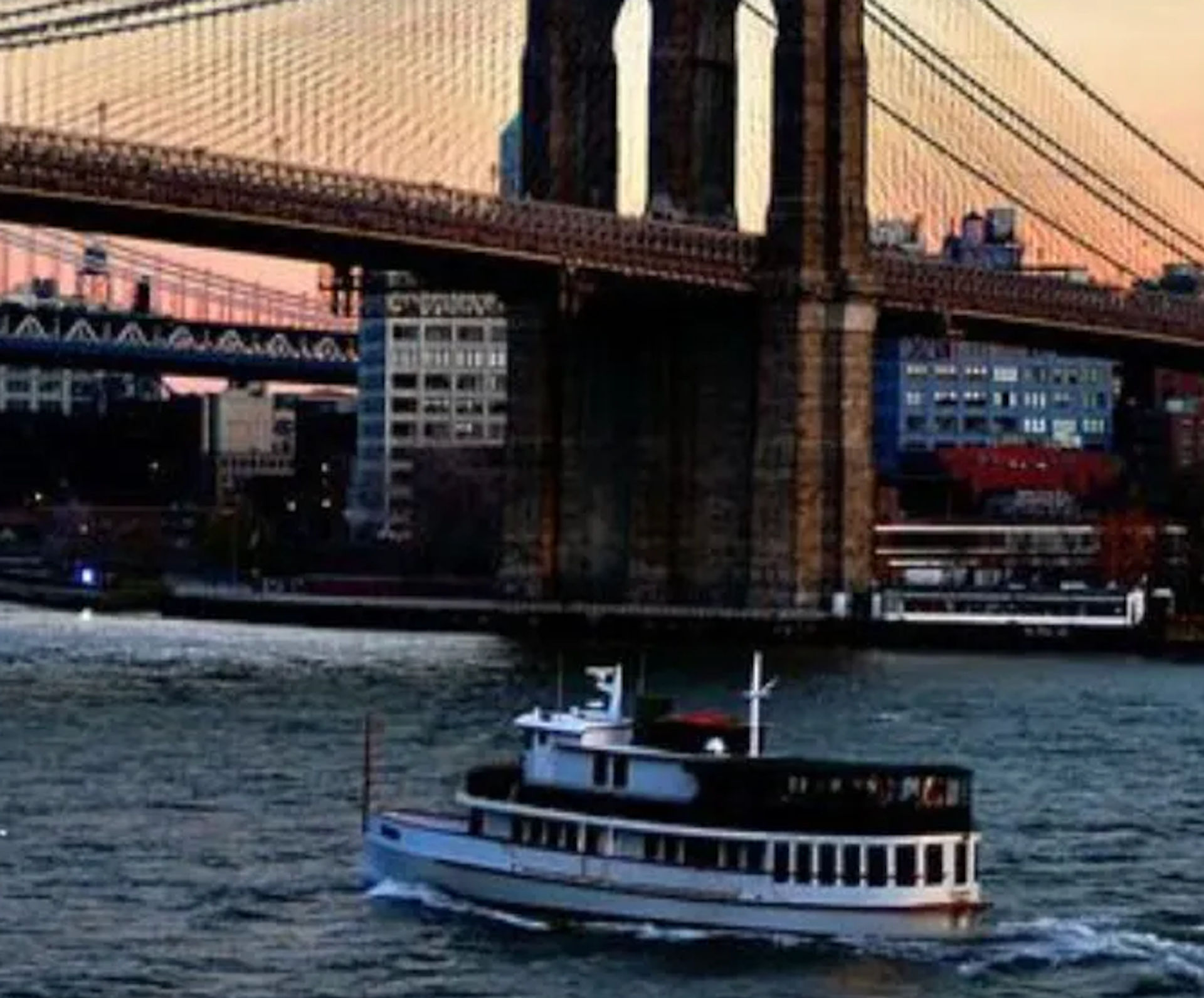 Sightseeing ferry chugging along the East River under the Brooklyn Bridge at sunset with Manhattan skyline in the background