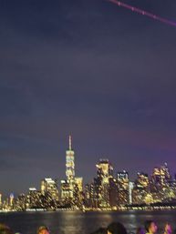 Night view of the New York City skyline across the Hudson River, illuminated skyscrapers with a glowing spire, water reflections and a blurred crowd in the foreground