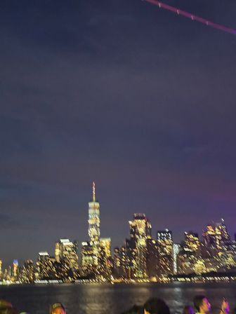Night view of the New York City skyline across the Hudson River, illuminated skyscrapers with a glowing spire, water reflections and a blurred crowd in the foreground
