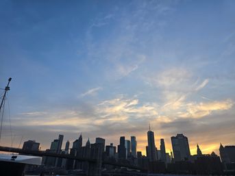Lower Manhattan skyline and Brooklyn Bridge silhouette at sunset, glowing golden clouds against a soft blue sky.