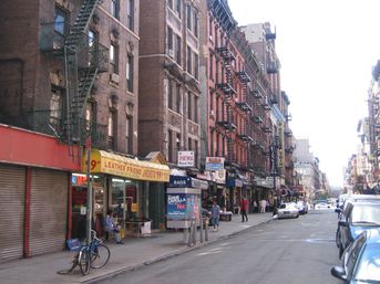 Sunlit New York City street scene with rows of brick tenement buildings and metal fire escapes, ground‑floor retail shops with awnings, parked cars, a bicycle and pedestrians.
