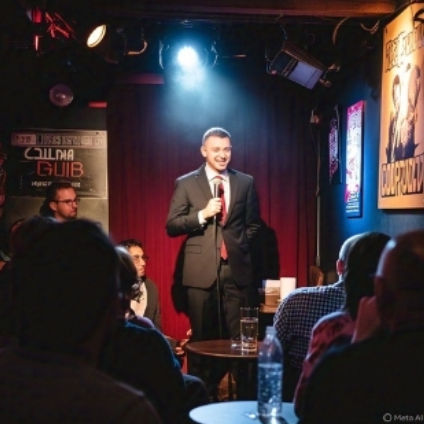 Stand-up comedian in a suit smiling under a spotlight on a small comedy club stage with a red curtain backdrop and a seated audience at tables.