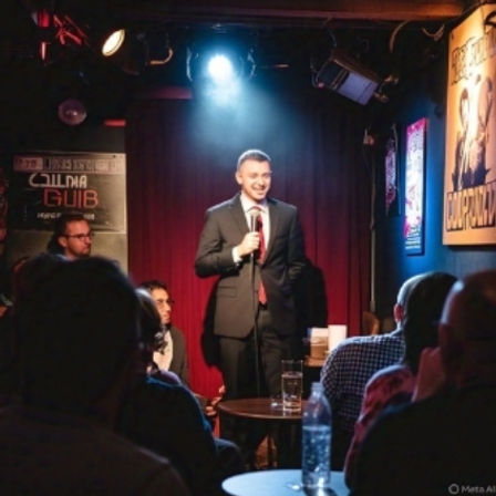 Stand-up comedian in a suit smiling under a spotlight on a small comedy club stage with a red curtain backdrop and a seated audience at tables.