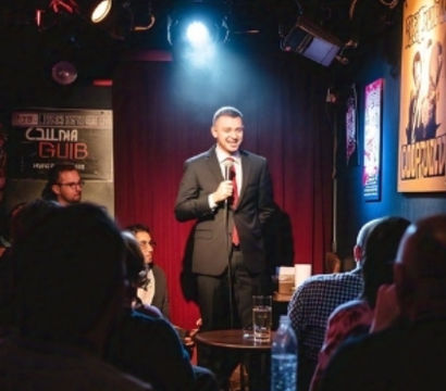 Stand-up comedian in a suit smiling under a spotlight on a small comedy club stage with a red curtain backdrop and a seated audience at tables.