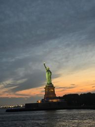 Illuminated Statue of Liberty on Liberty Island at sunset, torch glowing against dramatic clouds over New York Harbor