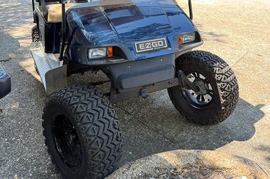 Lifted blue EZGO golf cart with oversized off-road tires and diamond-plate side step parked on a sunlit residential driveway