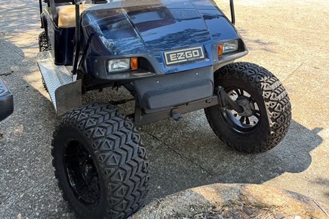 Lifted blue EZGO golf cart with oversized off-road tires and diamond-plate side step parked on a sunlit residential driveway