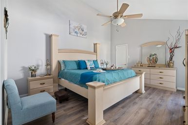 Bright, airy master bedroom with teal quilted bedding, light wood bed frame and dresser, ceiling fan, blue accent chair, nightstand with lamp and flowers, and wood-look flooring — a cozy, coastal-inspired retreat.