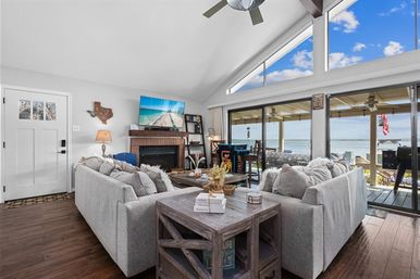 Sunny waterfront open-plan living room with vaulted ceiling and fan, paired gray sectionals around a wooden coffee table and fireplace with TV, hardwood floors and wide sliding glass doors opening to a covered deck and water view.