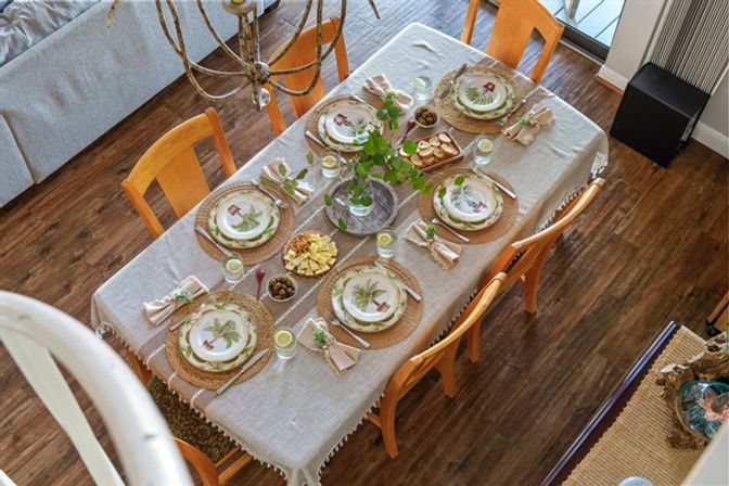Overhead view of a cozy home dining table set for six with a linen tablecloth, wicker placemats, botanical plates, folded napkins, glasses, snack bowls and a leafy centerpiece on hardwood floors.