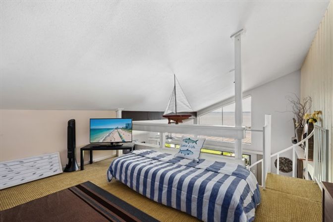 Coastal-themed loft bedroom with low sloped ceiling, blue-and-white striped daybed and fish pillow, hanging model sailboat, TV showing a beach scene, and large slanted windows with ocean view.