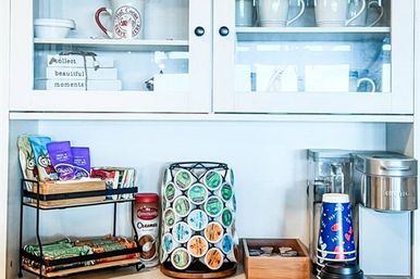 Bright white kitchen coffee station on countertop with glass-front cabinets, single-serve coffee maker, rotating coffee pod carousel, stacked snacks, creamer jar and disposable cups