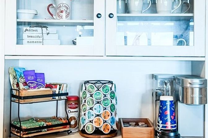 Bright white kitchen coffee station on countertop with glass-front cabinets, single-serve coffee maker, rotating coffee pod carousel, stacked snacks, creamer jar and disposable cups