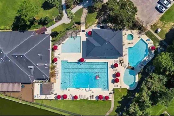 Aerial view of a suburban community pool complex with a large rectangular lap pool, smaller freeform pools, red umbrellas on the deck, clubhouse, green lawn and nearby parking