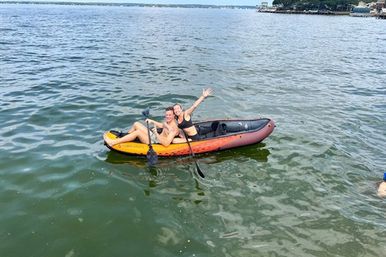 Two people in swimsuits paddling and waving from an orange inflatable kayak on a calm lake with a distant tree-lined shoreline — summer water recreation