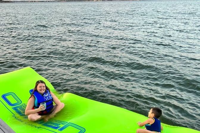 Smiling woman holding a drink and a child in life jackets relaxing on a bright green inflatable water mat floating on a calm lake near a dock.