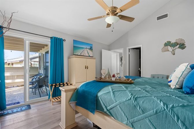 Sunlit coastal-style bedroom with teal quilted bedding, blue curtains, light wood furniture, ceiling fan and sliding glass door opening to a small patio with outdoor seating