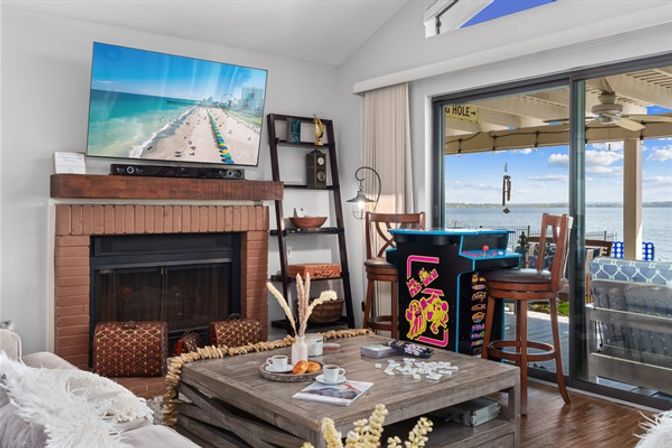 Cozy waterfront living room with brick fireplace and wall TV showing a beach, rustic coffee table with coffee and pastries, retro arcade game cabinet and bar stools by sliding glass doors opening to a sunny patio and calm water view.