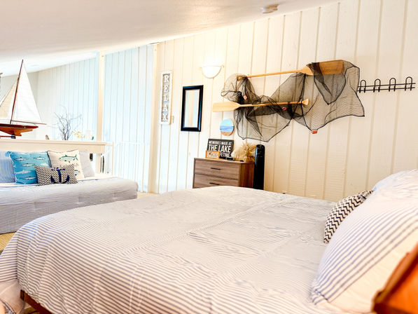 Relaxing coastal-themed loft bedroom with striped bedding, nautical throw pillows, model sailboat, wall-mounted oars draped with a fishing net and bright beach-cottage decor