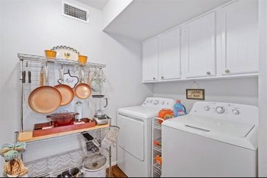 Bright residential laundry room with white top-load washer and dryer beneath white cabinets, metal shelving storing copper pans, blender, cookware and laundry detergent in a compact organized utility space.