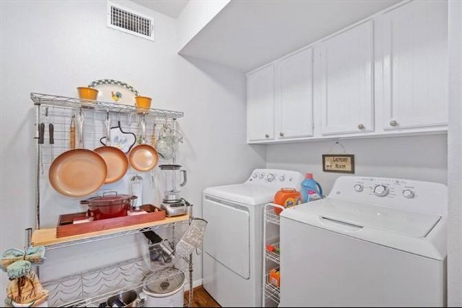Bright residential laundry room with white top-load washer and dryer beneath white cabinets, metal shelving storing copper pans, blender, cookware and laundry detergent in a compact organized utility space.