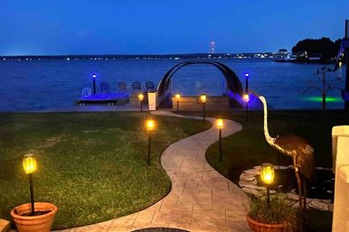 Twilight lakefront scene with a winding stone pathway lined by glowing lanterns leading across a lawn to an arched, blue-lit dock, with a metal heron sculpture and potted plants in the foreground.