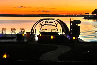 Serene lake sunset with orange sky reflecting on calm water, waterfront dock with curved boat lift, empty chairs and glowing pathway lights