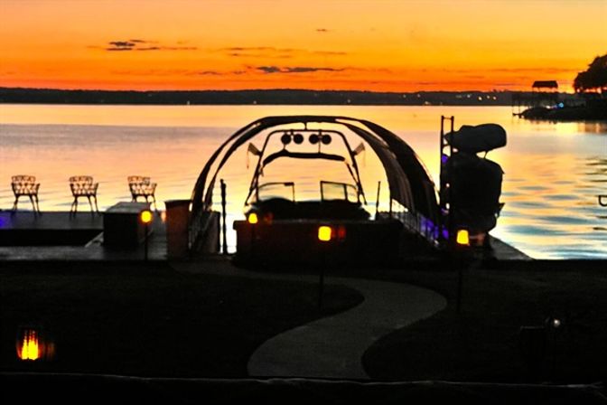 Serene lake sunset with orange sky reflecting on calm water, waterfront dock with curved boat lift, empty chairs and glowing pathway lights
