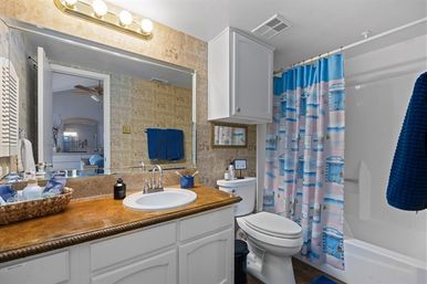 Coastal-style residential bathroom with brown vanity countertop, white sink and cabinets, large mirror with overhead lights, toilet, bathtub with blue-and-pink patterned shower curtain and coordinating blue towels.
