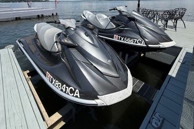 Two charcoal-gray jet skis with light-gray seats on lift platforms at a wooden lake dock in Texas, calm water and patio chairs nearby.