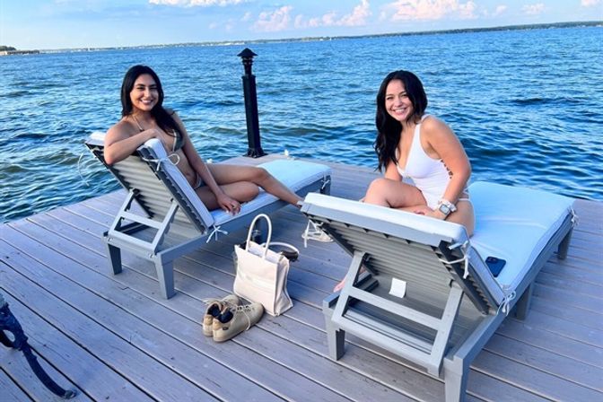 Two women in swimsuits lounging on adjustable chairs on a sunny lakeside dock, calm blue water and clear sky, beach bag and shoes on the wooden deck